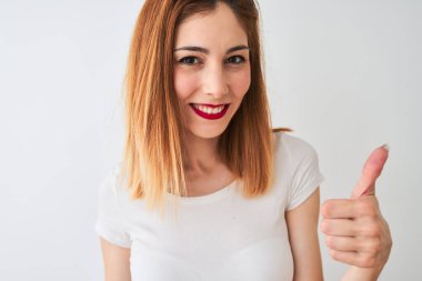 Güzel kızıl saçlı kadın, günlük tişört giyiyor, beyaz arka plan üzerinde duruyor, gülümsüyor, gülümsüyor, işaret veriyor, parmaklarıyla başparmağını kaldırıyor, mükemmel bir işaret.