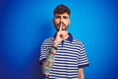 İzole edilmiş mavi arka planda duran çizgili polo giyen genç adam sessiz olmak istiyor. Sessizlik ve gizli konsept.