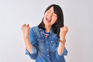 İzole edilmiş beyaz arka plan üzerine kot gömlek giyen genç, güzel Çinli bir kadın mutlu ve heyecanlı bir şekilde kollarını kaldırıp, gülümseyerek ve başarı için çığlık atarak kazanan bir jest yapıyor. Kutlama konsepti.