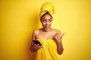 Afro kadın duş üstüne havlu giyiyor sarı arka plan üzerinde smatrphone kullanıyor ve gülümseyen bir gülümsemeyle baş parmağıyla işaret ediyor.