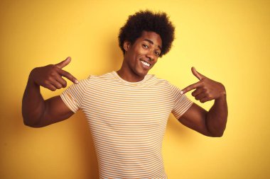 Afro saçlı, çizgili tişörtlü, izole edilmiş sarı arka planda duran, kendine güvenen, yüzünde gülümseme olan, kendini gururlu ve mutlu gösteren Amerikalı bir adam..