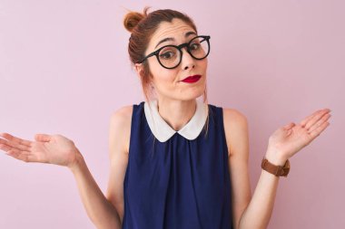 Saçları örgülü, zarif bir elbise ve gözlük takan kızıl saçlı bir kadın soyutlanmış pembe bir arka plan üzerine cahil ve elleri ve kolları havada kafası karışmış bir ifade takınıyor. Şüphe kavramı.
