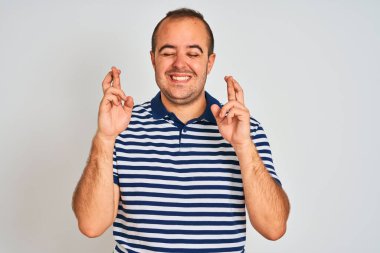 İzole edilmiş beyaz arka plan üzerinde duran sıradan çizgili polo giyen genç adam umut ve gözler kapalı gülümseyerek el sallıyor. Şans ve batıl inanç..