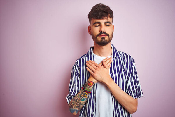 Young man with tattoo wearing striped shirt standing over isolated pink background smiling with hands on chest with closed eyes and grateful gesture on face. Health concept.