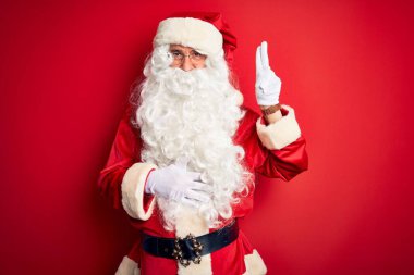 Orta yaşlı, yakışıklı, Noel Baba kostümü giymiş, izole edilmiş kırmızı arka planda durmuş gülümseyen, elleri göğsünde ve parmakları havada, sadakat yemini eden bir adam.