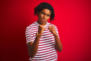 Siyah çizgili tişörtlü genç afro adam izole edilmiş kırmızı arka planda duruyor. Yumruk savunma hareketleriyle dövüşmeye hazır, kızgın ve üzgün yüzlü, sorundan korkan.