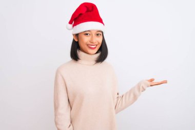 Noel Baba şapkası takan genç, güzel Çinli bir kadın izole edilmiş beyaz arka planda mutlu bir şekilde gülümsüyor ve avuç içi kameraya bakıyor..