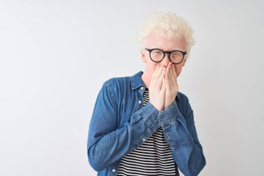 Genç albino sarışın adam kot gömlek ve gözlük takıyor beyaz arka planda gülüyor ve utanıyor kıkırdıyor ağızları ellerle, dedikoduyla ve skandallarla örtüyor.