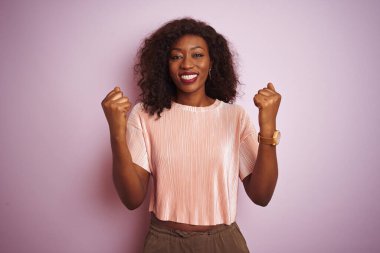 Genç Afro-Amerikan kadın tişört giyip pembe arka planda dikiliyor. Şaşırmış ve şaşırmış bir şekilde kolları açık bir şekilde başarıyı kutluyorlar. Kazanan konsept.