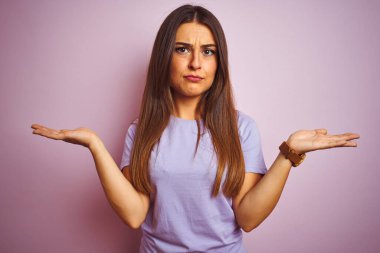 İzole edilmiş pembe arka plan üzerinde duran sıradan tişört giyen genç ve güzel bir kadın. Kolları ve elleri havada, kafası karışmış bir ifade. Şüphe kavramı.