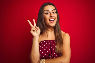 İzole edilmiş kırmızı arka plan üzerinde sıradan tişörtler giyen genç ve güzel bir kadın. Kameraya bakıp gülümsüyor ve zafer işareti yapıyor. İki numara..