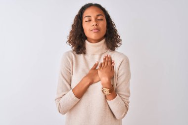 Balıkçı yaka kazak giyen genç Brezilyalı kadın izole edilmiş beyaz arka plan üzerinde duruyor elleri göğsünde, gözleri kapalı ve yüzünde minnet dolu bir jest ile gülümsüyor. Sağlık kavramı.