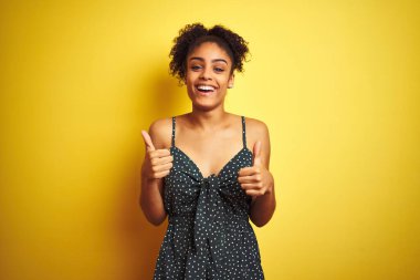 Afrika kökenli Amerikalı bir kadın, izole edilmiş sarı arka plan işareti üzerine günlük yeşil elbise giyip el hareketleriyle olumlu jest yapıyor, gülümsüyor ve mutlu. Neşeli ifade ve kazanan jest.
