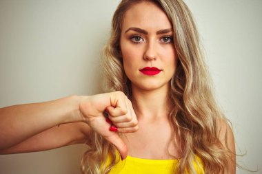 Sarı tişörtlü genç bir kadın öfkeli yüzüyle izole edilmiş beyaz bir arka plan üzerinde duruyor. Negatif işaretler baş parmakları aşağıda, reddedilme konsepti reddedilmişliği gösteriyor.