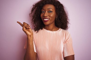 Genç Afro-Amerikan kadın tişört giyiyor pembe arka planda duruyor yüzünde kocaman bir gülümseme, eli ve parmağı kameraya bakıyor..