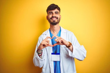 Dövmeli genç doktor, izole edilmiş sarı arka planda duruyor. Aşkla gülümsüyor. Elleriyle kalp sembolü ve şekli gösteriyor. Romantik konsept.