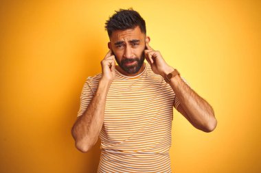 İzole edilmiş sarı arka planda duran tişört giyen genç Hintli adam yüksek sesli müzik sesi için kulakları parmaklarıyla kapatıyor. Sağır kavramı.