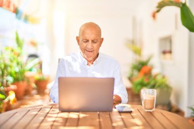 Kıdemli yakışıklı adam mutlu ve kendinden emin gülümsüyor. Terasta dizüstü bilgisayar kullanarak oturmak