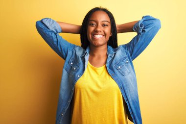 Genç Afro-Amerikalı kadın kot gömlek giyiyor, izole edilmiş sarı arka planda duruyor, rahatlıyor ve esniyor, kolları ve elleri başının arkasında mutlu bir şekilde gülümsüyor.