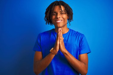 Rastalı tişörtlü Afro-Amerikan bir adam izole edilmiş mavi arka planda duruyor ellerini birleştirip af diliyor kendinden emin bir şekilde gülümsüyor..