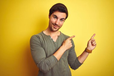 Sarı arka plan üzerinde duran rahat tişörtlü genç yakışıklı adam gülümsüyor ve kameraya bakıyor iki eliyle ve parmaklarıyla yan tarafa bakıyor..