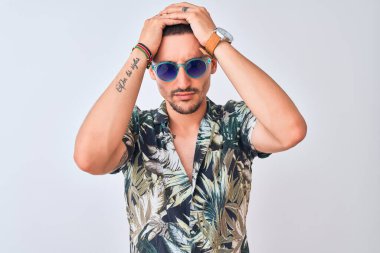 Hawaii süvari gömleği ve mavi güneş gözlüğü takan genç yakışıklı adam izole arka planda acı ve migren yüzünden çaresiz ve stresli bir baş ağrısı çekiyor. Ellerini başının üstüne koy..