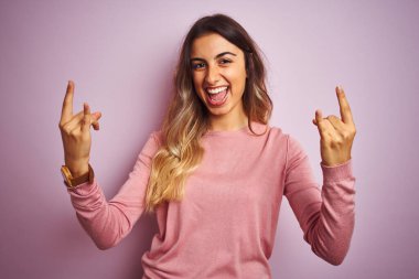 Pembe izole edilmiş arka planda kazak giyen genç ve güzel bir kadın çılgın bir ifadeyle el kaldırarak rock sembolü yapıyor. Müzik yıldızı. Ağır konsept.