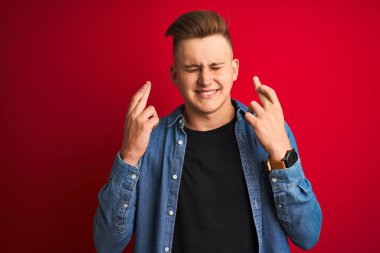 İzole edilmiş kırmızı arka plan üzerinde duran kot gömlek giyen yakışıklı genç adam umut ve gözlerin kapalı bir şekilde gülümseyerek işaret ediyor. Şans ve batıl inanç..