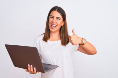 Güzel genç bir kadın bilgisayarını beyaz arka plan üzerinde kullanarak mutlu bir el hareketi yapıyor. Kameraya bakıp başarıyı gösteren ifade onaylanıyor.