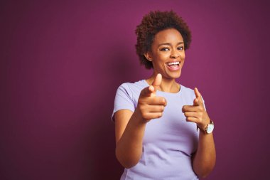 İzole edilmiş mor arka plan üzerinde saçı olan güzel Afro-Amerikalı kadın mutlu ve komik yüzlü parmaklarını kameraya doğrultuyor. İyi enerji ve titreşimler.