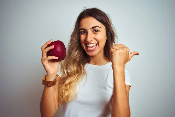 若いです美しいです女性食べる赤いリンゴ以上グレー隔離された背景指すと見せますとともに親指で側まで幸せな顔笑顔 — ストック写真