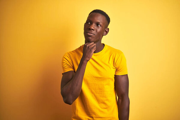 Young african american man wearing casual t-shirt standing over isolated yellow background with hand on chin thinking about question, pensive expression. Smiling with thoughtful face. Doubt concept.