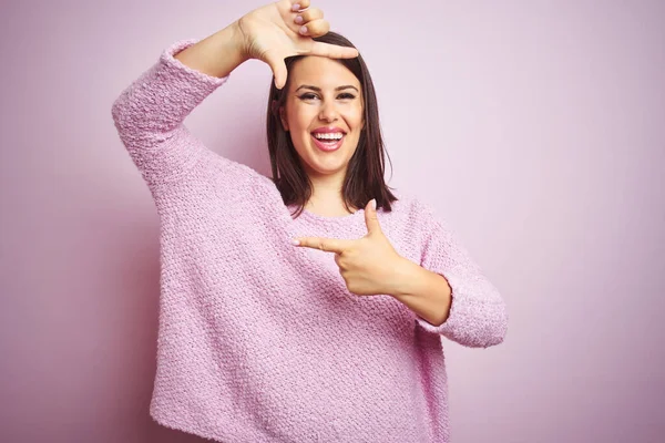 ピンクの孤立した背景にセーターを着た若い美しいブルネットの女性は 幸せな顔をした手と指でフレームを作り笑顔 創造性と写真の概念 — ストック写真