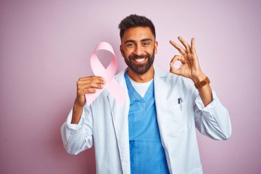 Kanser kurdelesi tutan genç Hintli doktor izole edilmiş pembe arka planda duruyor. Parmaklarıyla tamam işareti yapıyor. Mükemmel bir sembol.