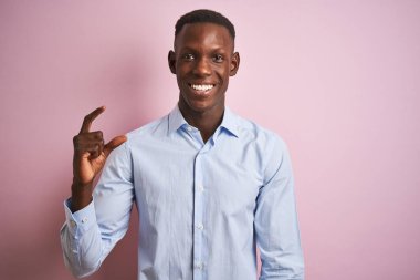 İzole edilmiş pembe arka plan üzerinde duran mavi şık gömlekli Afro-Amerikan bir adam gülümsüyor ve kendine güvenen el hareketleriyle küçük boy işareti yapıyor parmaklarıyla bakıyor ve kameraya bakıyor. Ölçü kavramı.