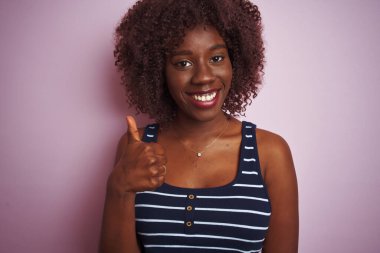 İzole edilmiş pembe arka planda duran çizgili tişört giyen Afrikalı genç bir kadın elleriyle mutlu bir jest yapıyor. Kameraya bakıp başarıyı gösteren ifade onaylanıyor.