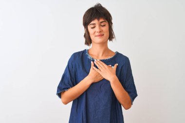 İzole edilmiş beyaz arka planda duran kot elbise giyen genç ve güzel bir kadın. Elleri göğsünde, gözleri kapalı ve yüzünde minnet dolu bir jest ile gülümsüyor. Sağlık kavramı.