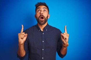 Soyutlanmış mavi arka plan üzerinde duran genç Hintli zarif adam şaşırmış ve şaşırmış görünüyor ve parmaklarıyla işaret ediyor ve kollarını kaldırıyor..