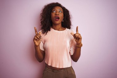 İzole pembe arka plan üzerinde duran t-shirt giyen genç Afro-Amerikan kadın şaşırttı ve yukarı bakarak ve parmakları ile işaret ve kolları kaldırdı şaşırttı.
