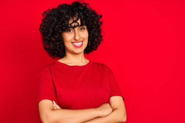 Kıvırcık saçlı genç Arap kadın izole edilmiş kırmızı arka plan üzerinde günlük tişört giyip kollarını kavuşturup kameraya bakarken gülümsüyor. Pozitif kişi.