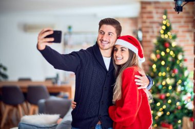 Mutlu ve kendinden emin gülümseyen genç bir çift. Ayakta durmak ve sarılmak, evdeki Noel ağacının etrafında kamerayla selfie çektirir.