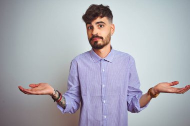 Mor gömlekli genç adam, soyutlanmış beyaz arka plan üzerinde duruyor. Kolları ve elleri havada, kafası karışmış bir ifade. Şüphe kavramı.