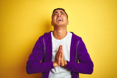 Mor eşofman üstü giyen genç Brezilyalı adam izole edilmiş sarı arka planda duruyor. Elleriyle yalvarıyor ve dua ediyor. Yüzünde umut ifadesi var. Af diliyor. Din kavramı.