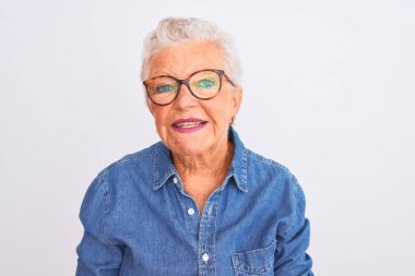Yaşlı, gri saçlı, kot gömlek ve gözlüklü bir kadın izole edilmiş beyaz arka planda ellerini birleştirip gülümseyerek gevşemiş ve neşeli gülümsüyor. Başarı ve iyimser