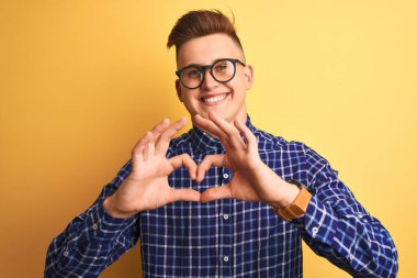 Sarı arka planda gözlüklü, gündelik tişört takan yakışıklı genç bir adam. Aşkla gülümsüyor. Elleriyle kalp sembolü ve şekli gösteriyor. Romantik konsept.