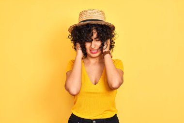 Kıvırcık saçlı, tişörtlü ve şapkalı genç bir Arap kadın izole edilmiş sarı arka planda acı ve migrenden dolayı çaresiz ve stresli bir baş ağrısı çekiyor. Ellerini başının üstüne koy..