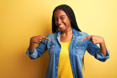Genç Afro-Amerikalı kadın kot gömlek giyiyor. İzole edilmiş sarı arka plan üzerinde duruyor. Yüzünde bir gülümsemeyle, gururlu ve mutlu bir şekilde kendini işaret ediyor..
