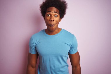 Afro saçlı, mavi tişörtlü, izole edilmiş pembe arka planda duran, dudaklı, çılgın ve komik bir jest yapan Afro-Amerikan bir adam. Komik ifade.