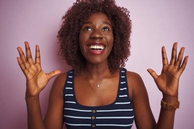 İzole edilmiş pembe arka planda duran çizgili tişört giyen Afrikalı genç bir kadın başarının çılgınlığını kollarını kaldırmış ve gözlerini kapatmış heyecanla haykırarak kutluyor. Kazanan konsept