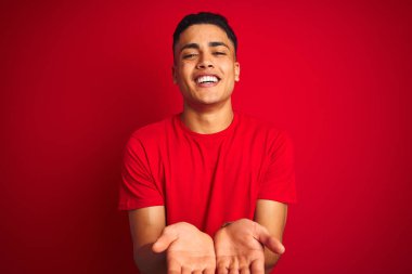 İzole edilmiş kırmızı arka plan üzerinde dikilen Brezilyalı genç adam avuçlarını birbirine uzatarak gülümsüyor ya da el hareketi yapıyor. Tutun ve koruyun